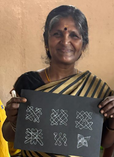An older participant smiling and holding up a black sheet covered in white kolam patterns she has drawn.